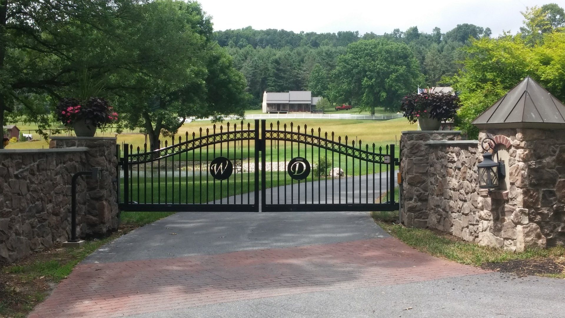 Black wrought-iron gates with stone pillars and urns, leading to a green field and a distant house.