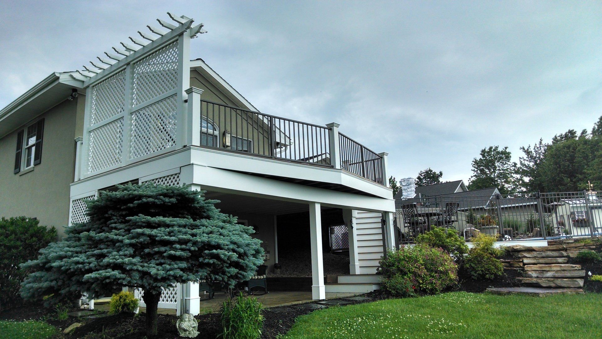 Two-story house with white deck and trellis. Green lawn with bushes and trees under a cloudy sky.