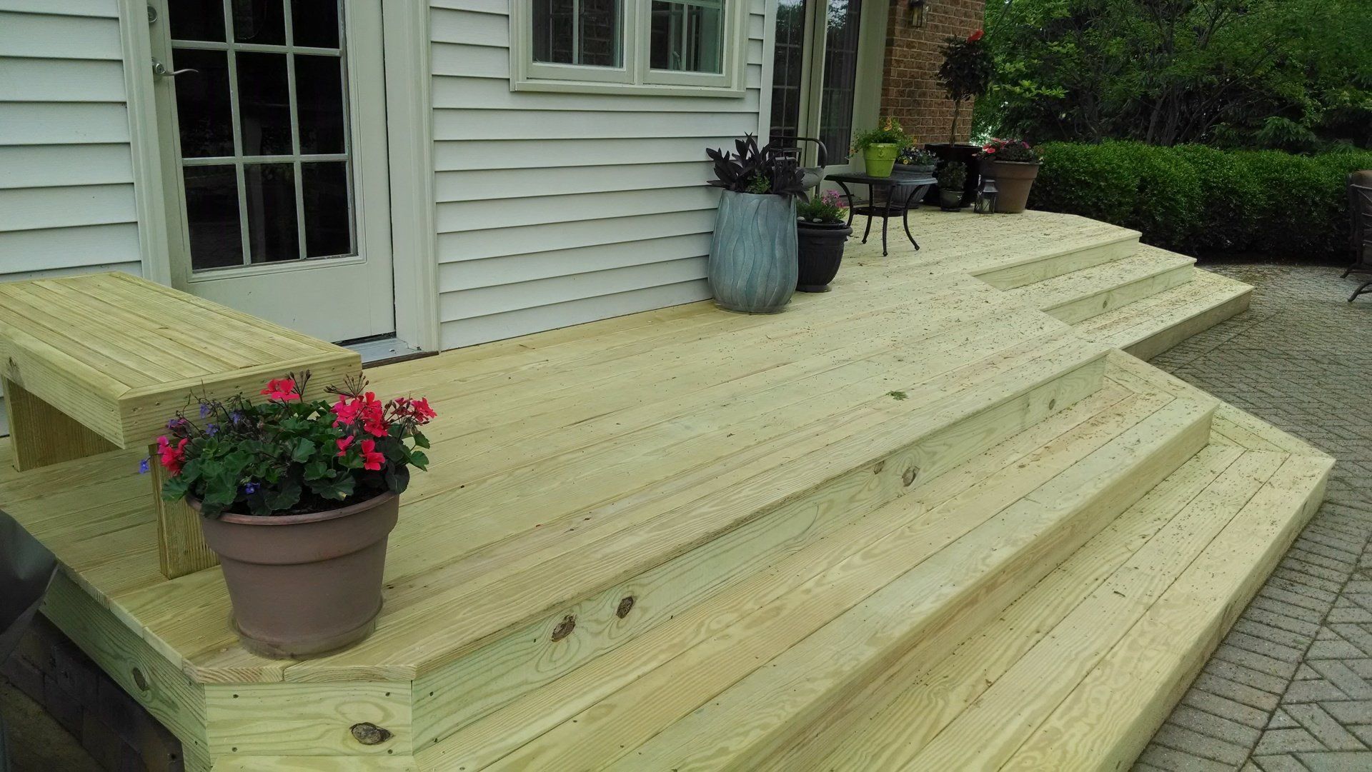 Wooden deck with steps and potted plants next to a white house with a door and window.
