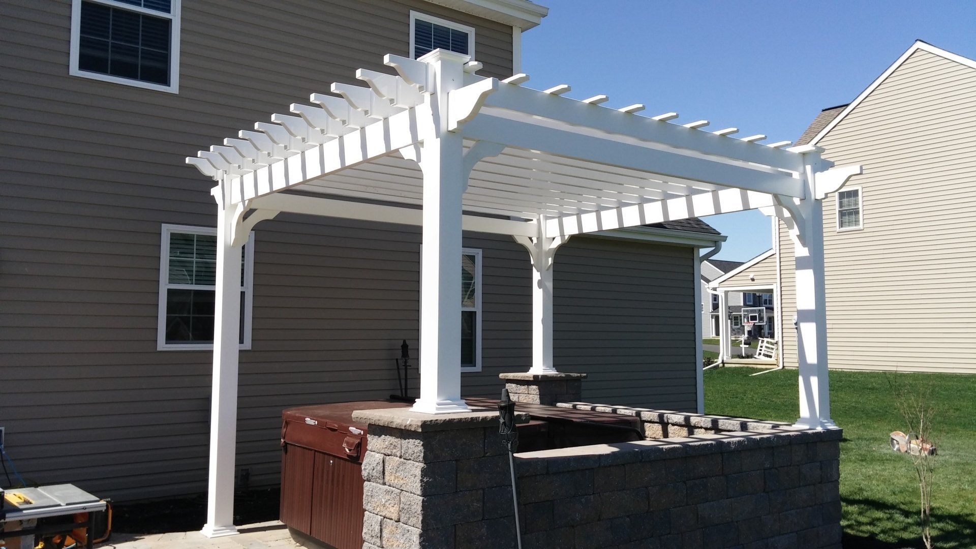 White pergola over a hot tub, set against a two-story beige house and sunny sky.