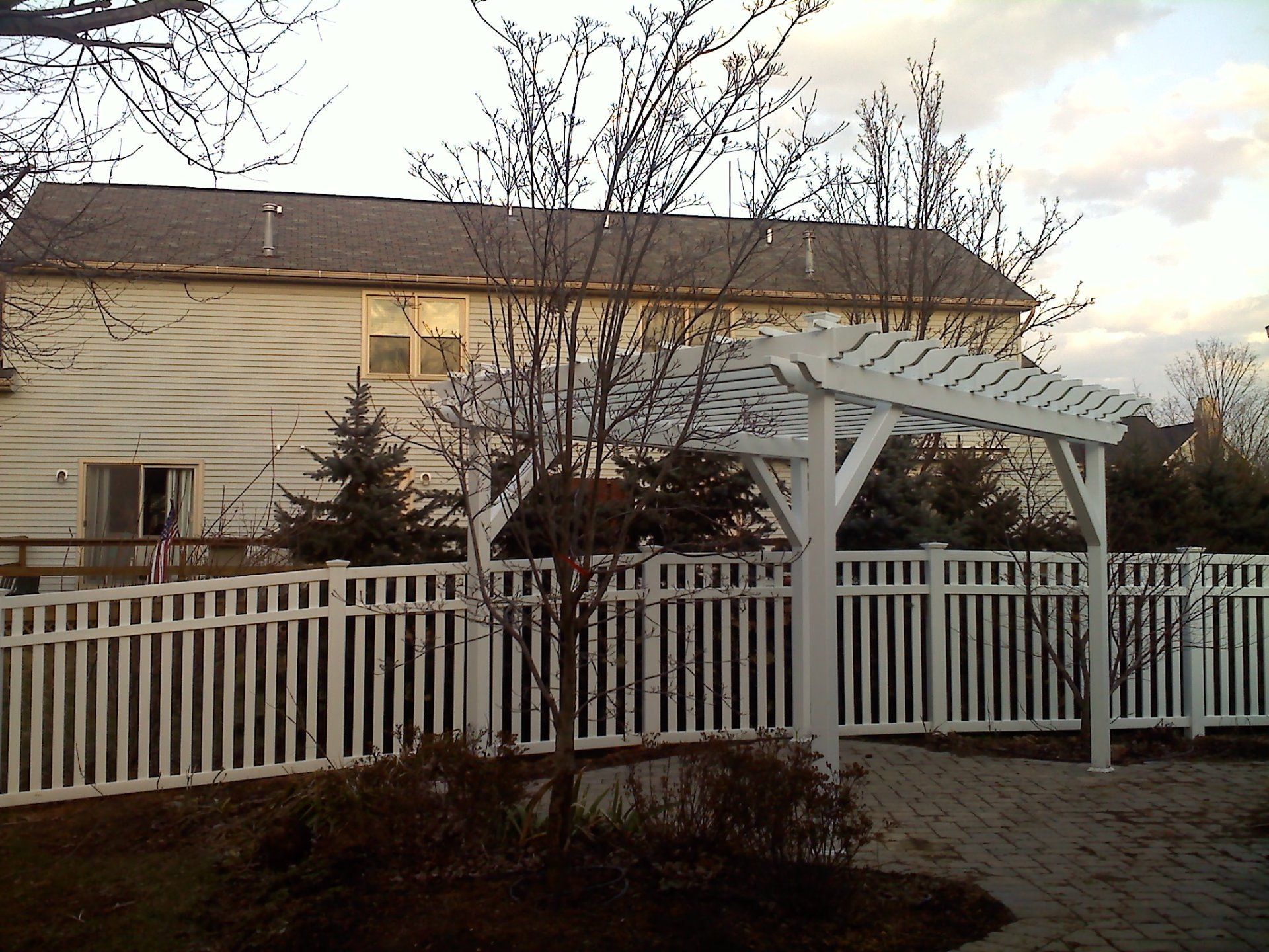 White picket fence and pergola in front of a light-colored house. Trees and a brick patio are visible.
