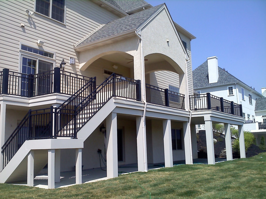 Two-story deck with black railings and stairs attached to a tan house with a grassy yard and blue sky.