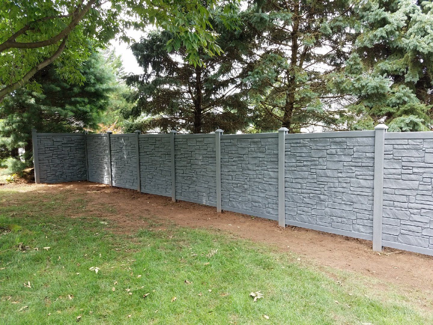 Gray stone-textured privacy fence in a grassy yard, with trees in the background.
