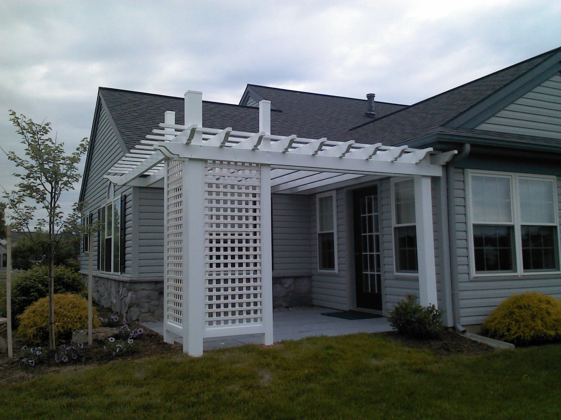 White pergola attached to a light gray house, over a patio, on a grassy lawn.
