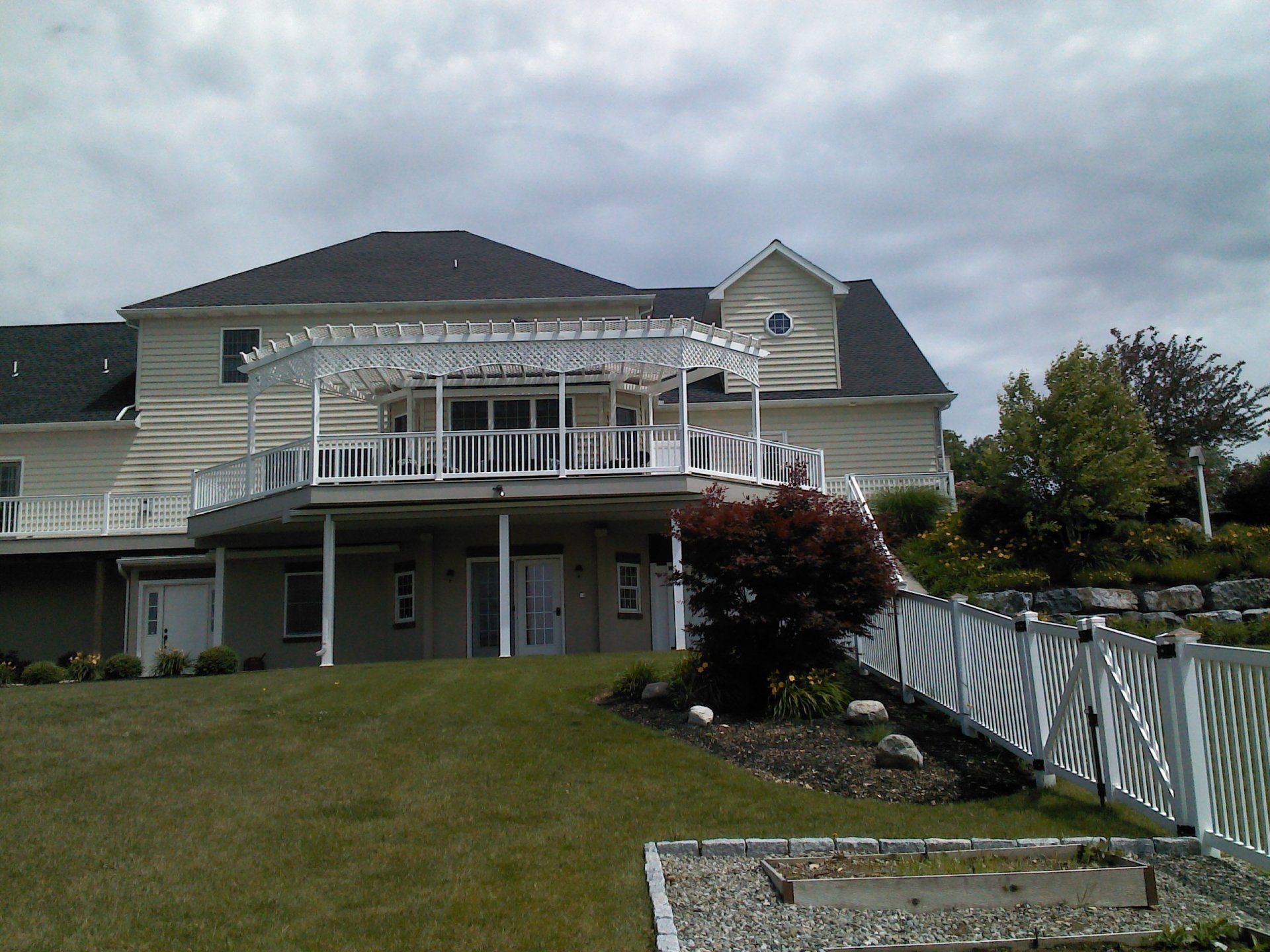 Large beige house with a wraparound deck and white pergola. A white fence is in the foreground, with a grass lawn.