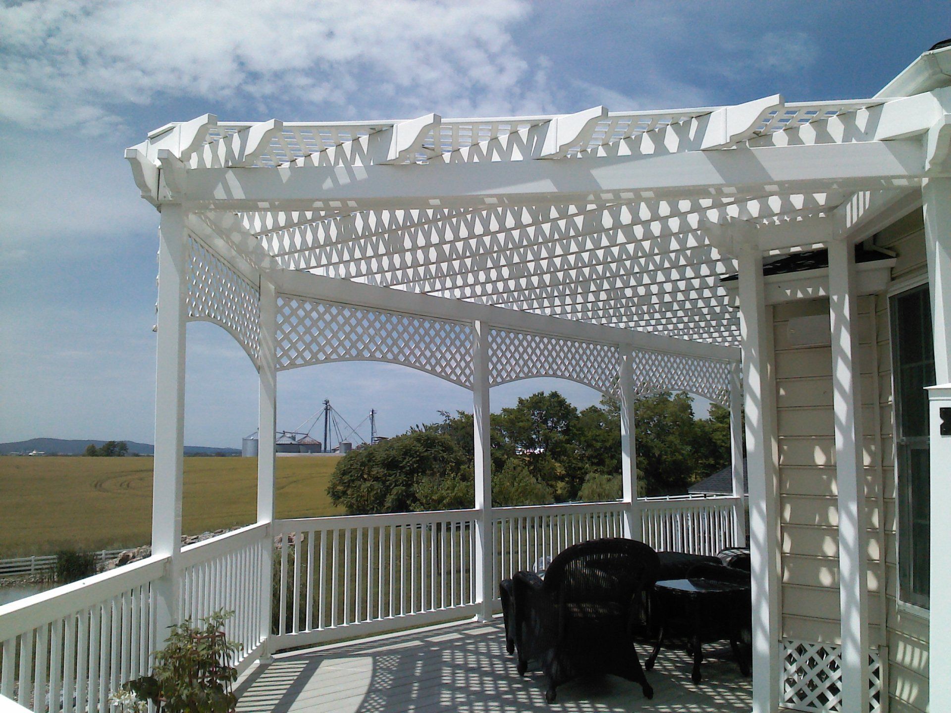 White latticed pergola on a balcony overlooking a field and distant structures.