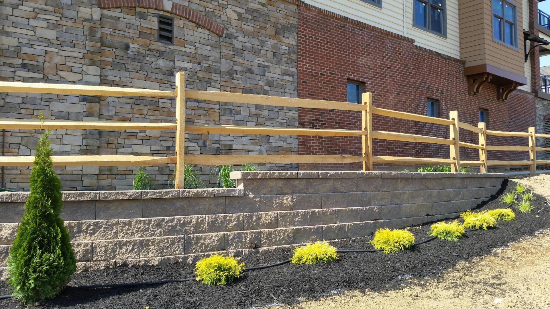 Wooden split-rail fence atop a retaining wall with shrubs. Building with stone and brick facade in background.
