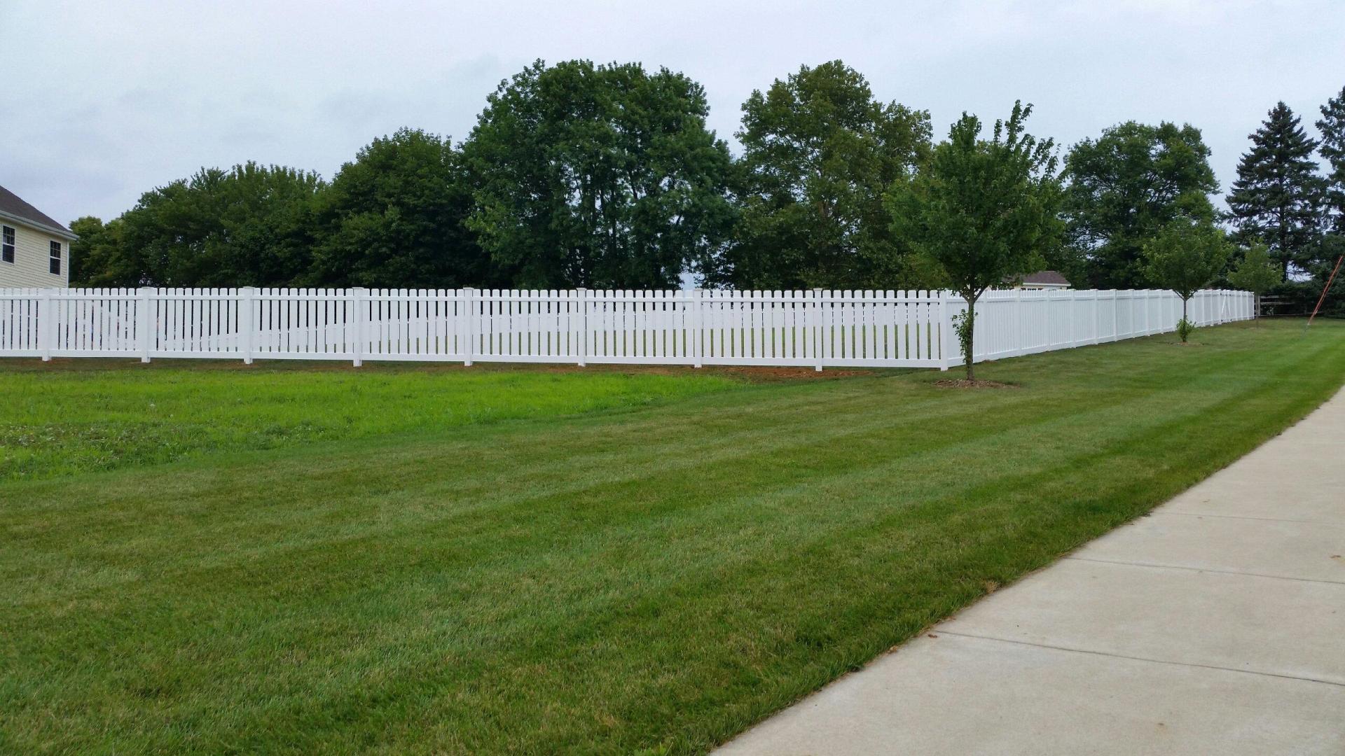 White picket fence bordering a grassy lawn, trees in background, sidewalk in foreground.