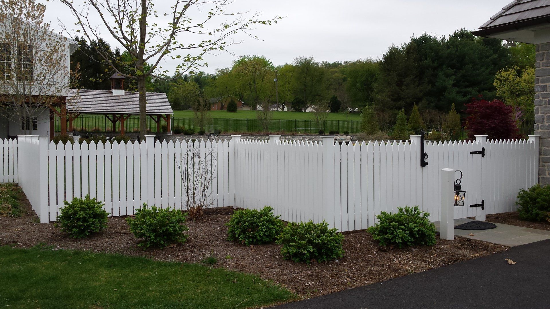 White picket fence bordering a yard with shrubs and a gate, overlooking a pond and trees.