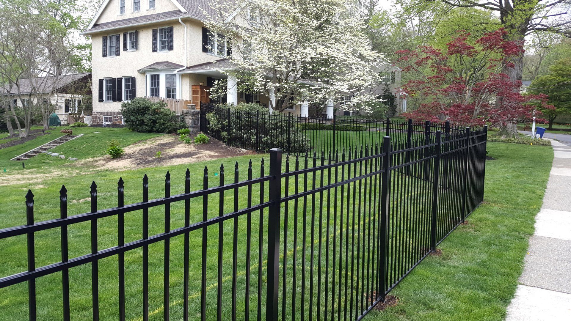 Black metal fence in front of a house with a green lawn and trees.
