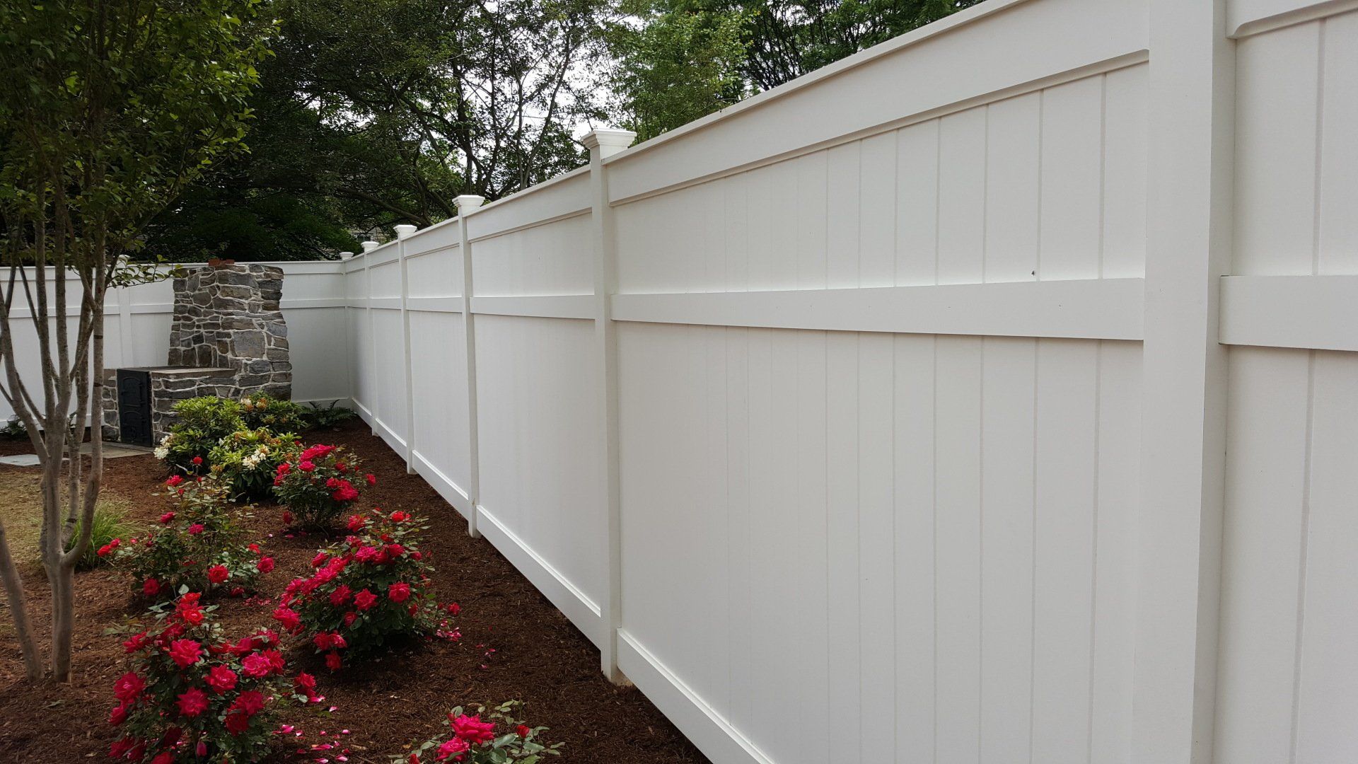 White privacy fence bordering a garden with red flowers and a tree.