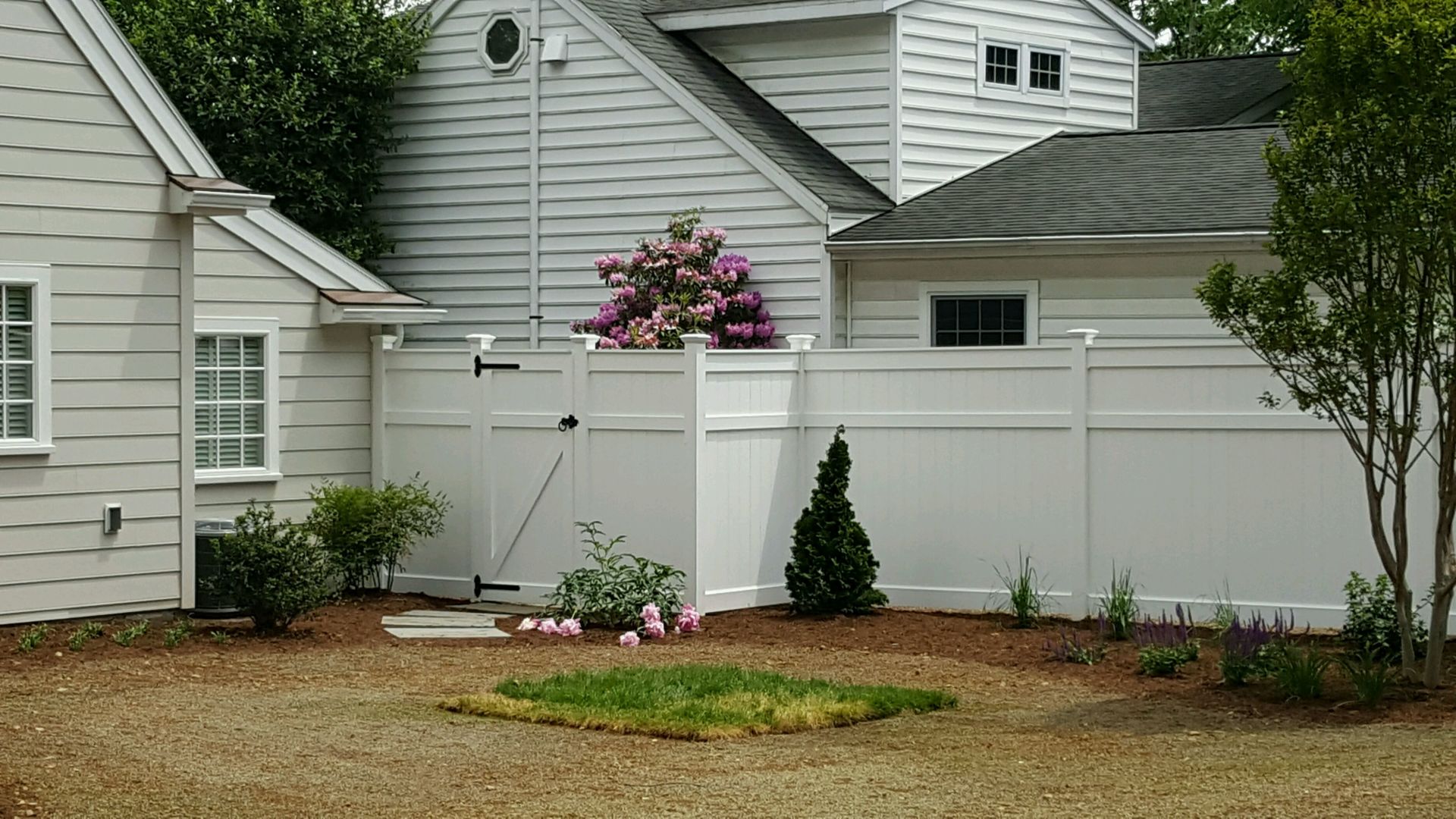 White vinyl fence encloses a backyard with small trees and bushes in front of a light-colored house.