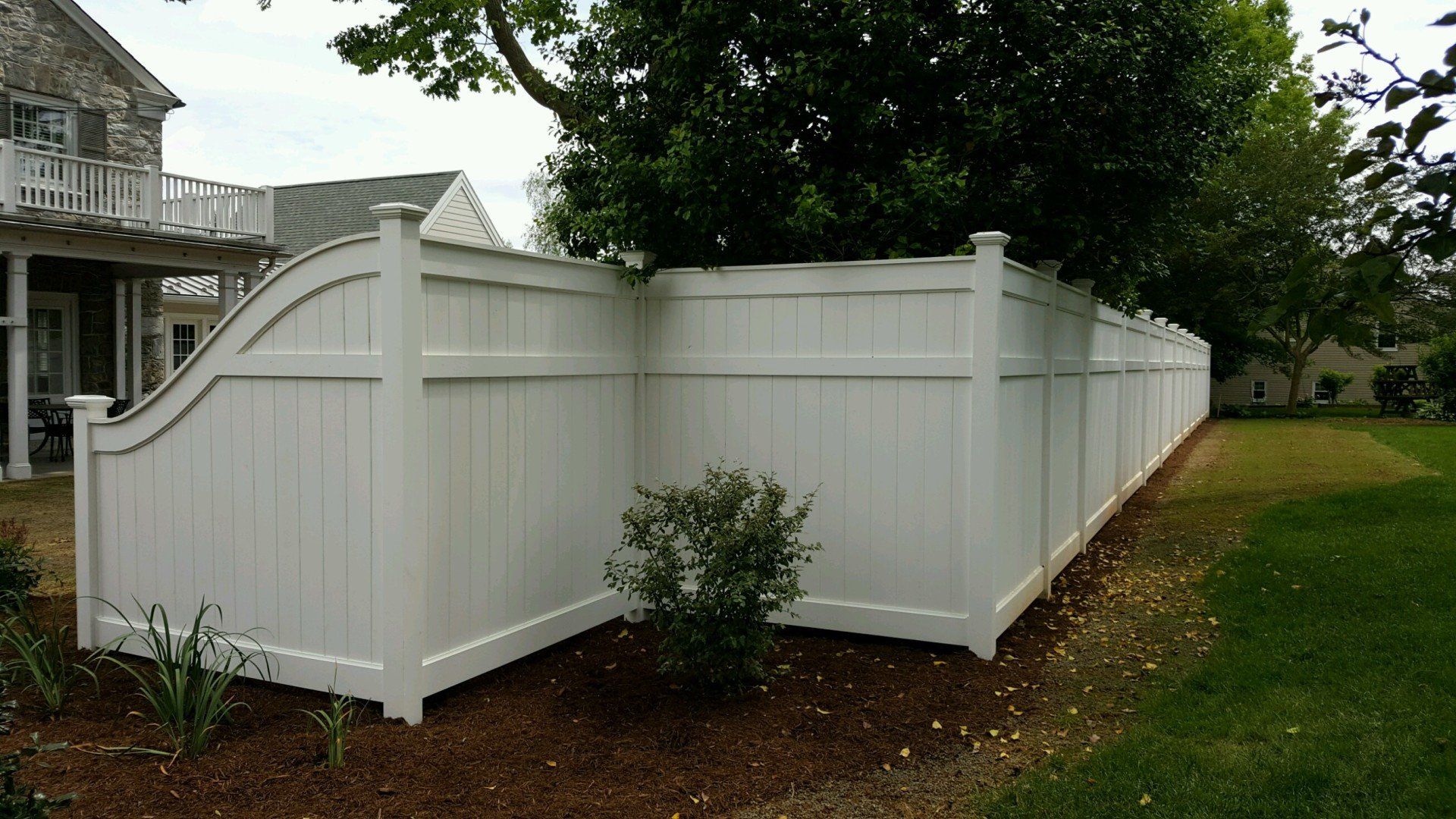 White vinyl privacy fence around a grassy yard, partially obscuring a house and tree.