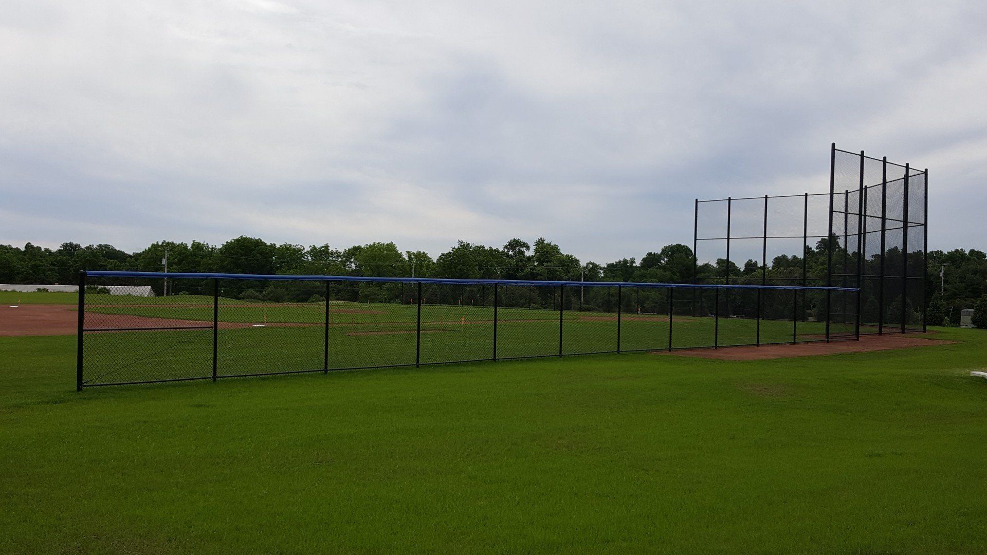 Baseball field with a long net, green grass, and cloudy sky.
