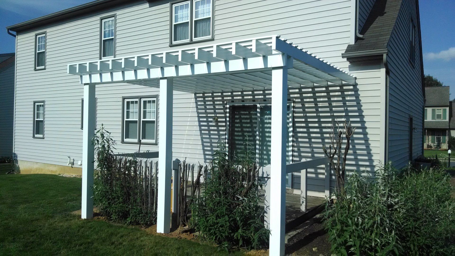 White pergola attached to a light gray house, casting shadows on the wall and plants below.