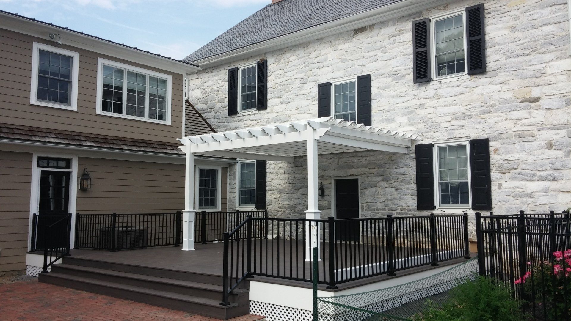 Exterior view of a two-story stone building with black shutters, a white pergola, and a brown deck.