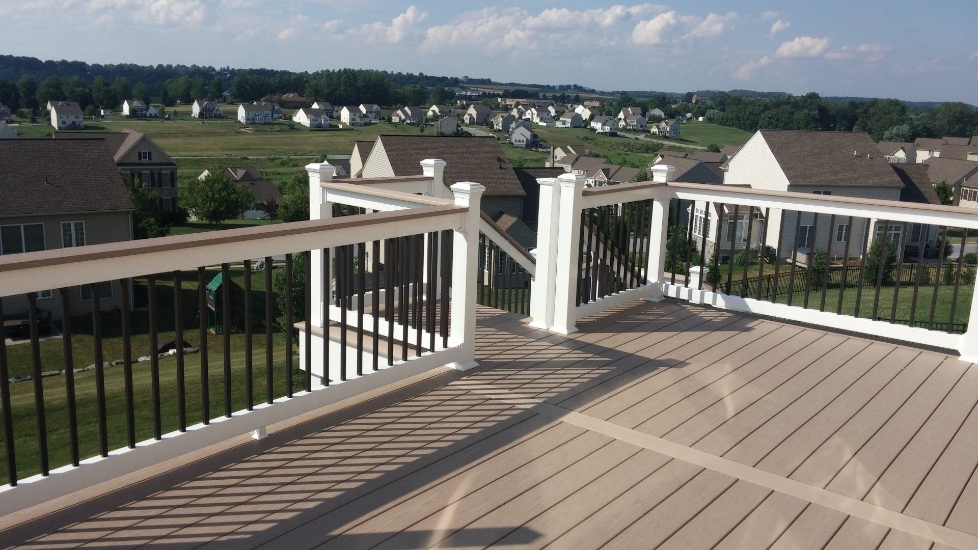 Wooden deck with white railing and black spindles, overlooking a suburban neighborhood on a sunny day.