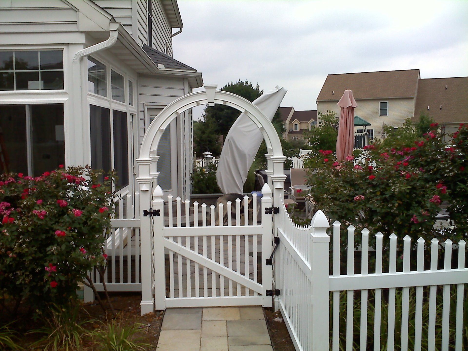 White picket fence with arched gate; roses in front, houses in the background.