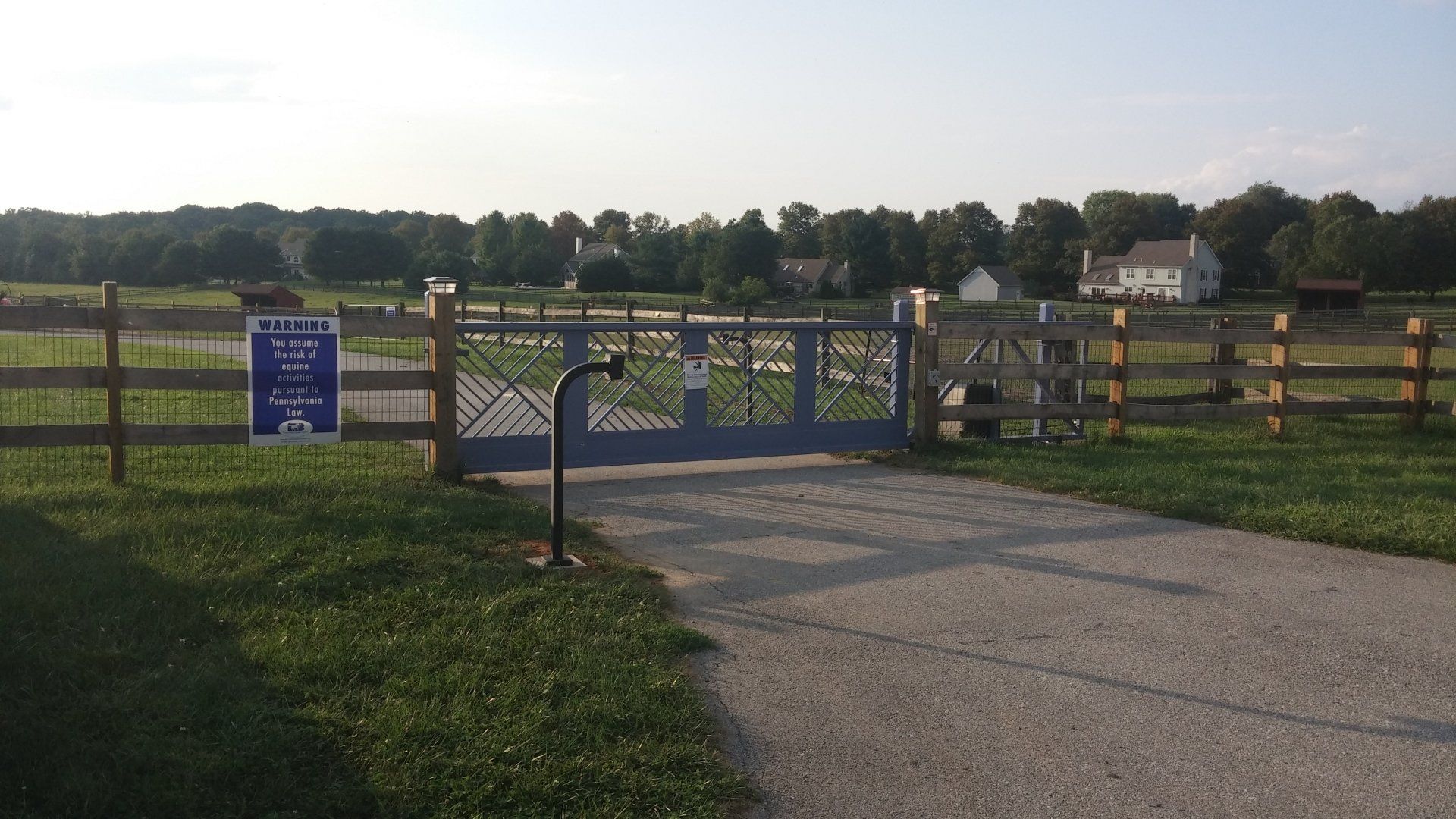Gate and driveway leading to a field with houses in the background. Blue gate, wooden fence, sign.