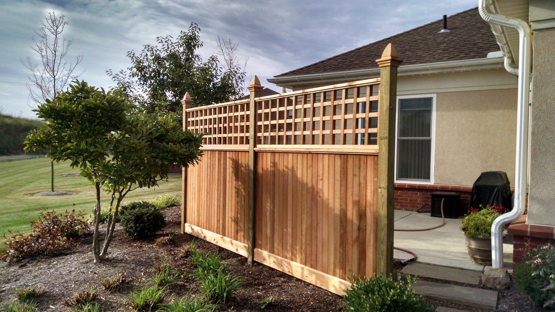Wooden fence with lattice top beside a house, next to a small tree and greenery.