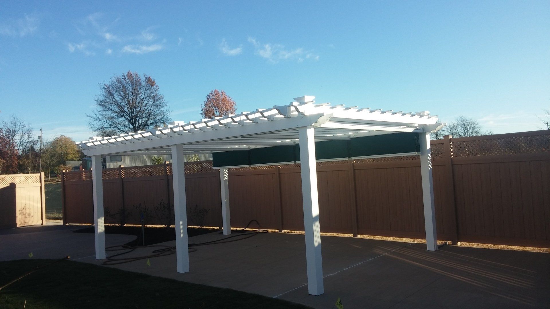 White pergola with green shade covers, set on a concrete patio, with a brown fence in the background.