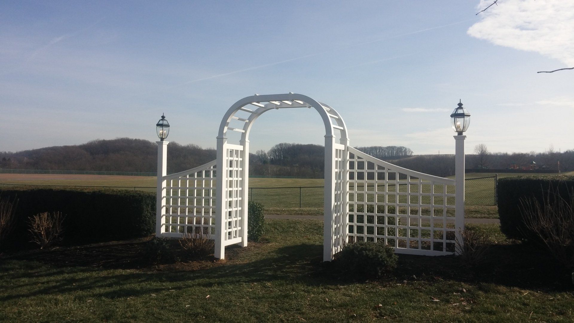 White arched trellis with lampposts opening to a field and treeline under a blue sky.