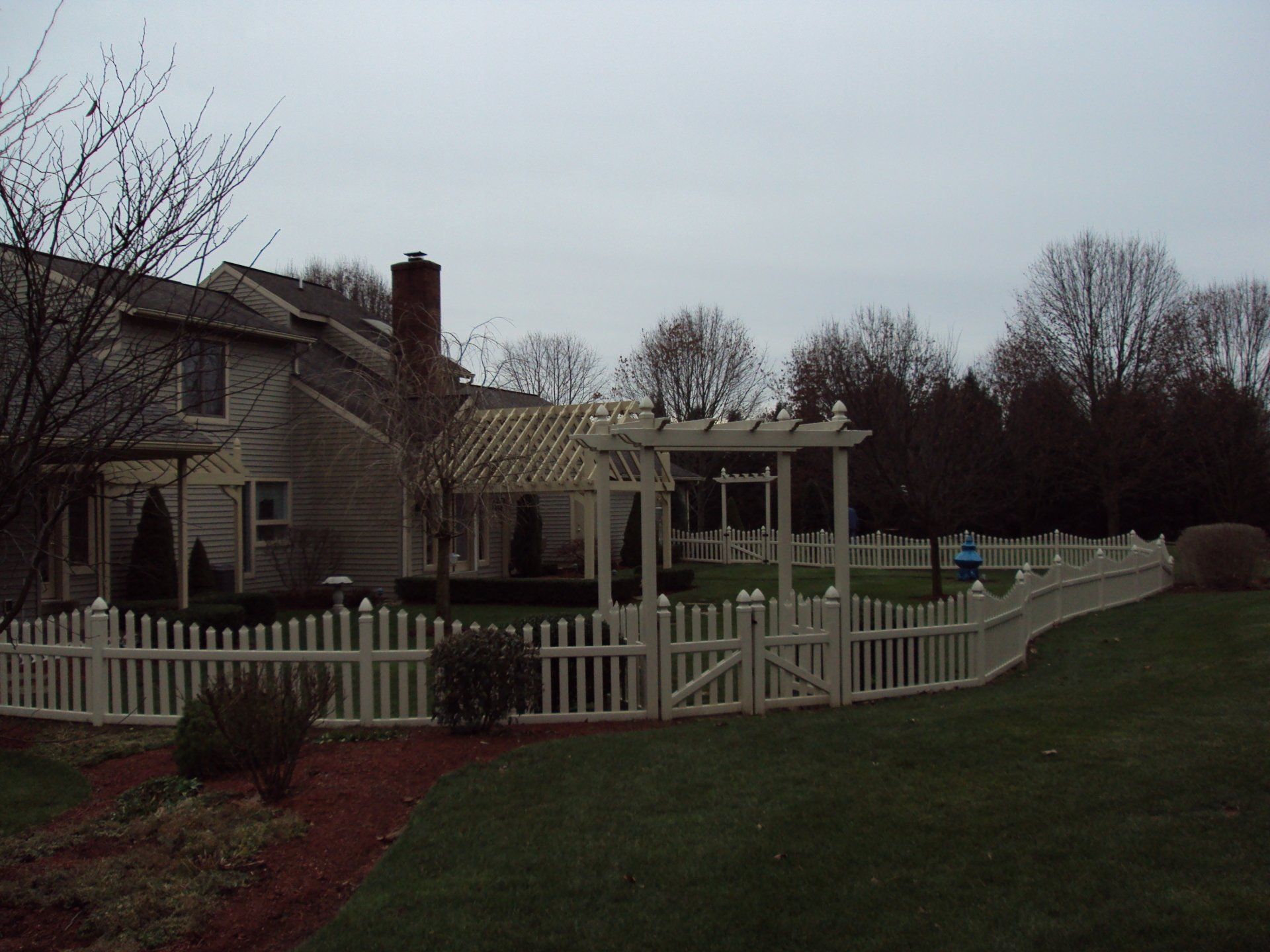 Beige picket fence surrounding a backyard with a house, pergola, and trees under a cloudy sky.