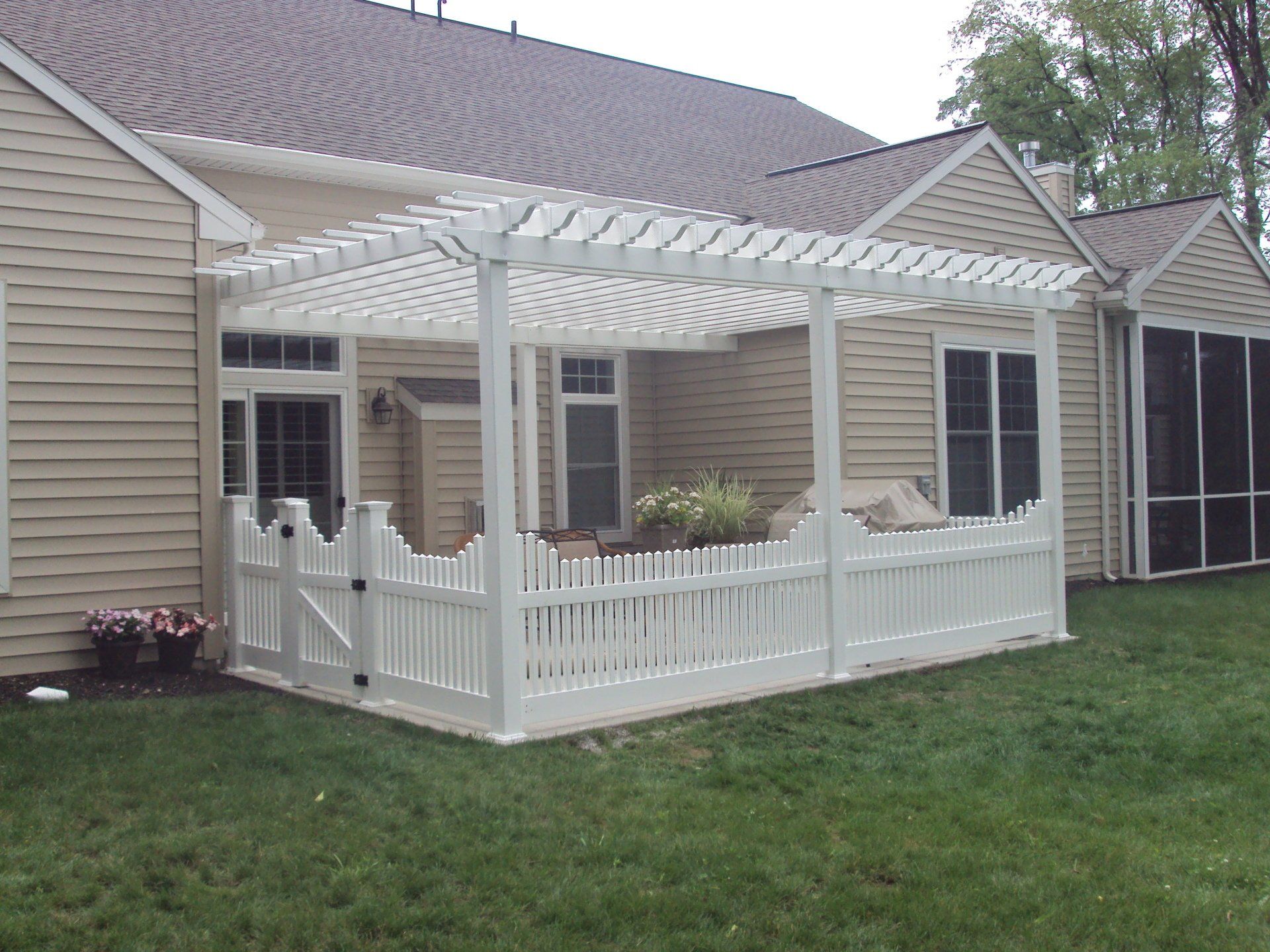 White pergola and picket fence on a patio attached to a beige house, green grass.