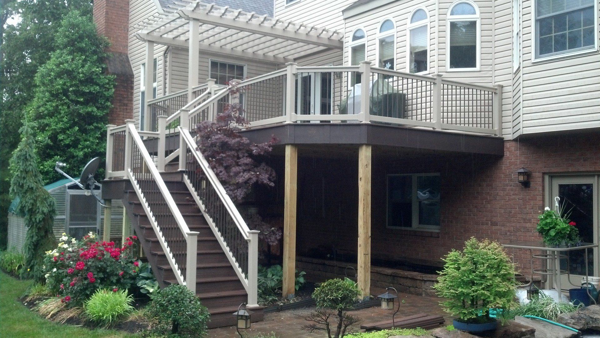 Multi-level brown deck with stairs, pergola, and light-colored railings attached to a brick house, surrounded by plants.