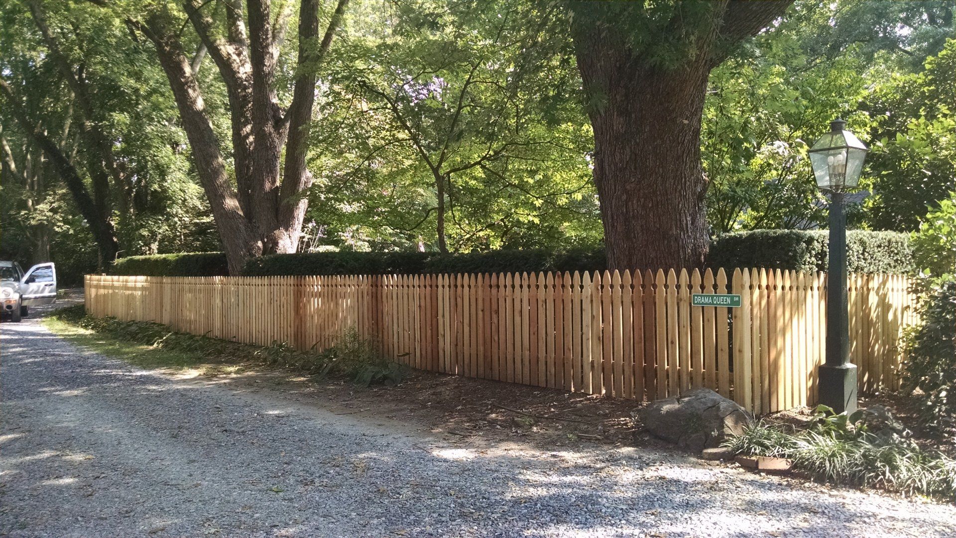 Wooden picket fence lines a gravel driveway, with trees in the background.