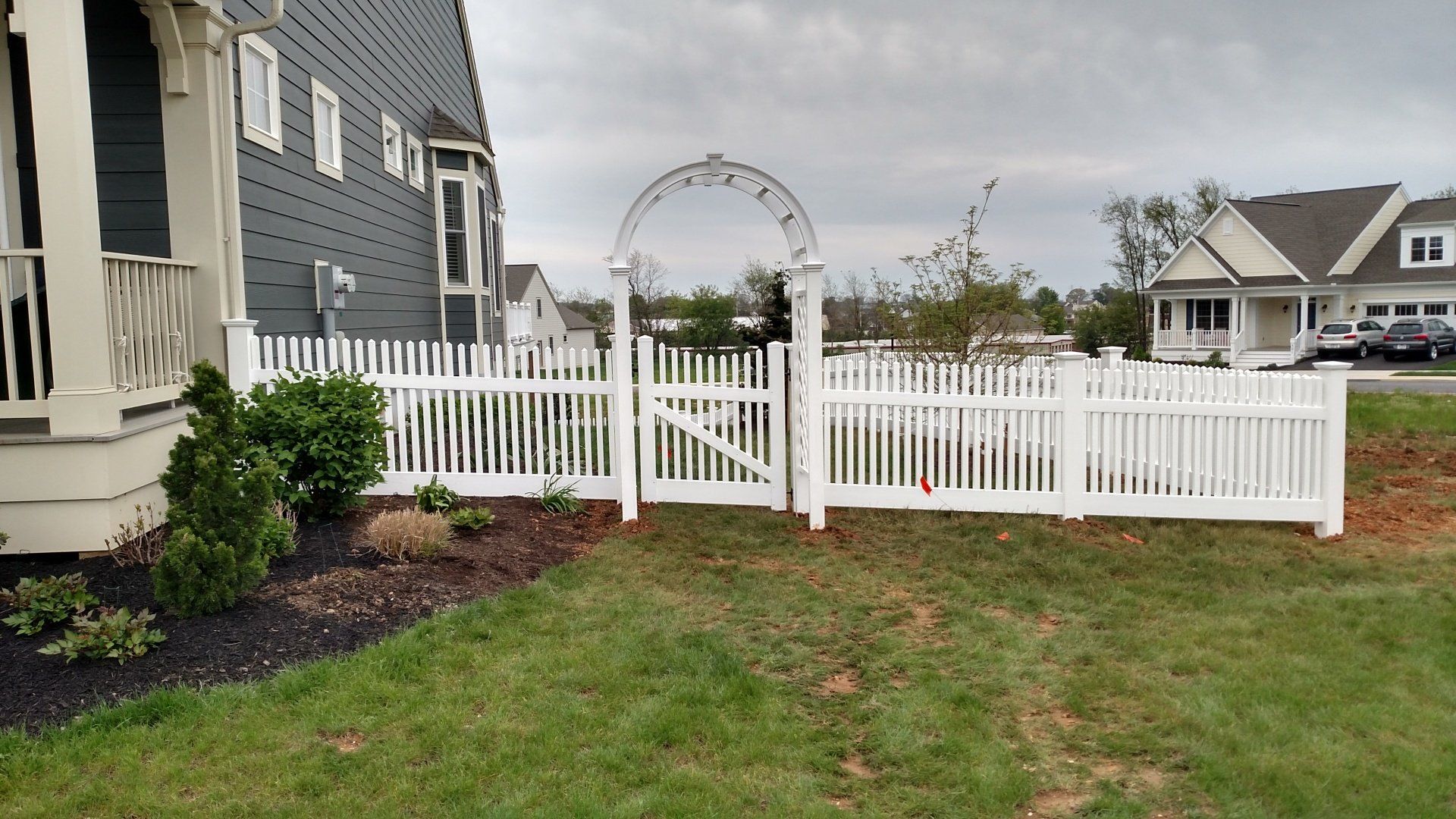White picket fence by Millcreek Fence & Decks with gate and arch, surrounding yard next to a gray house.