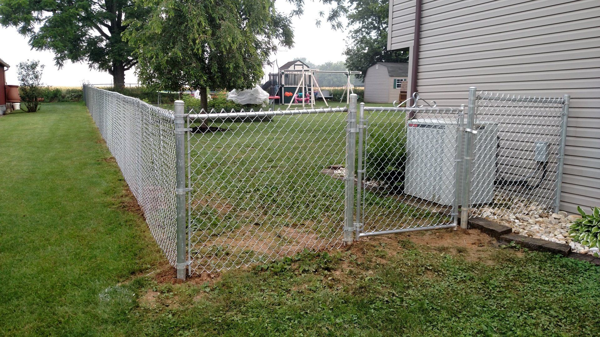 Chain-link fence in a backyard, with a gate near an air conditioning unit and house.