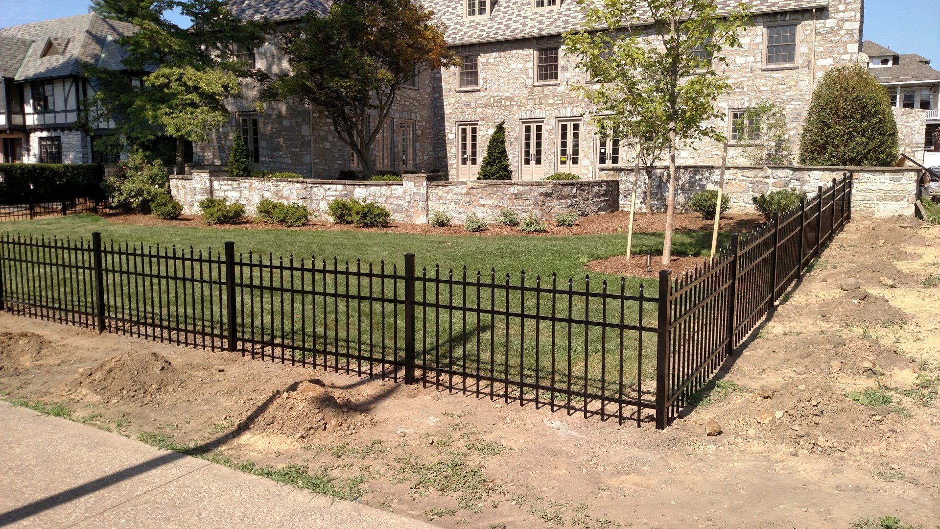 Black metal fence surrounds a grassy lawn, with a stone building in the background. Sunny day.