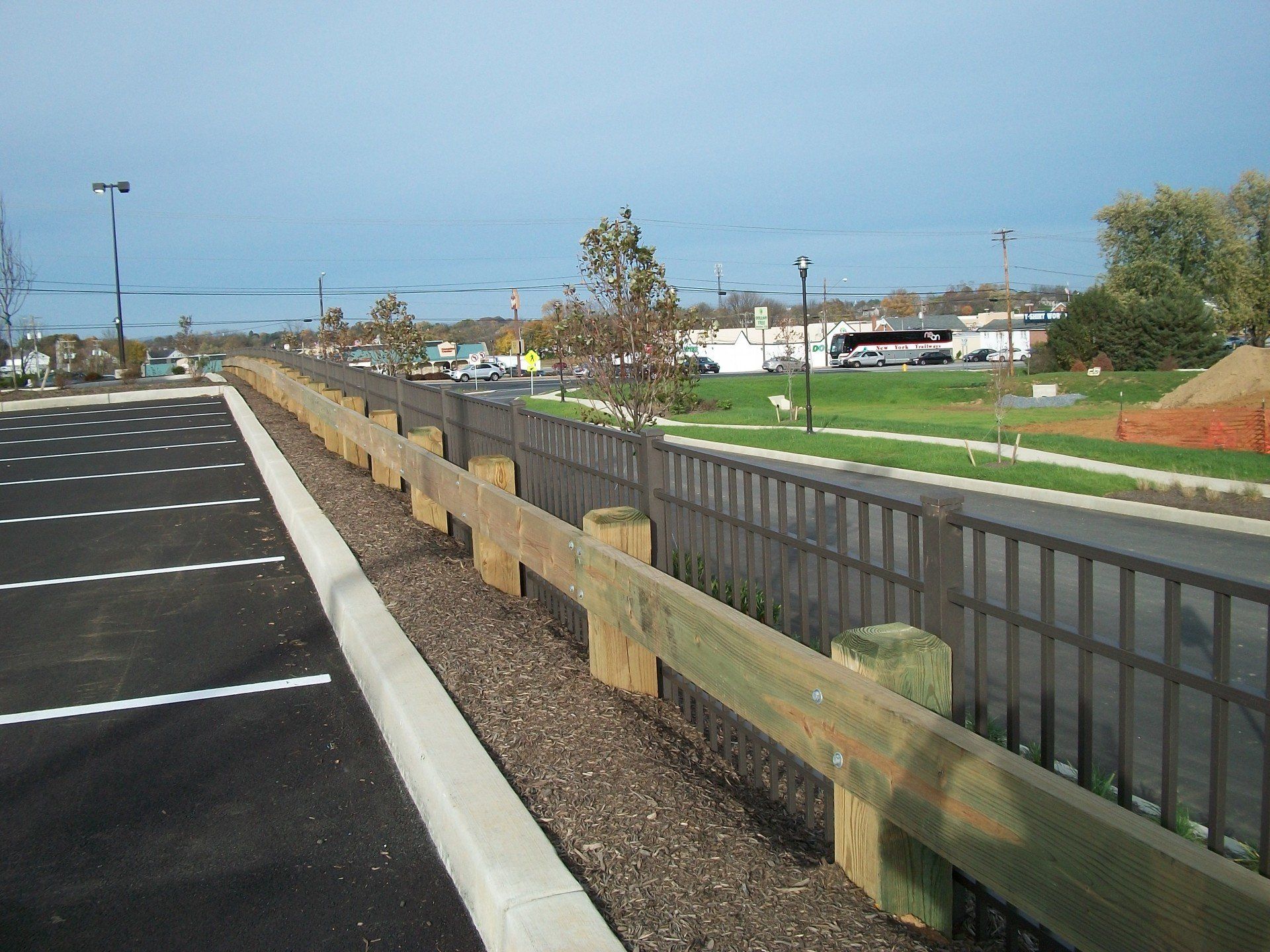 Wooden guardrail and metal fence alongside a road and parking lot.