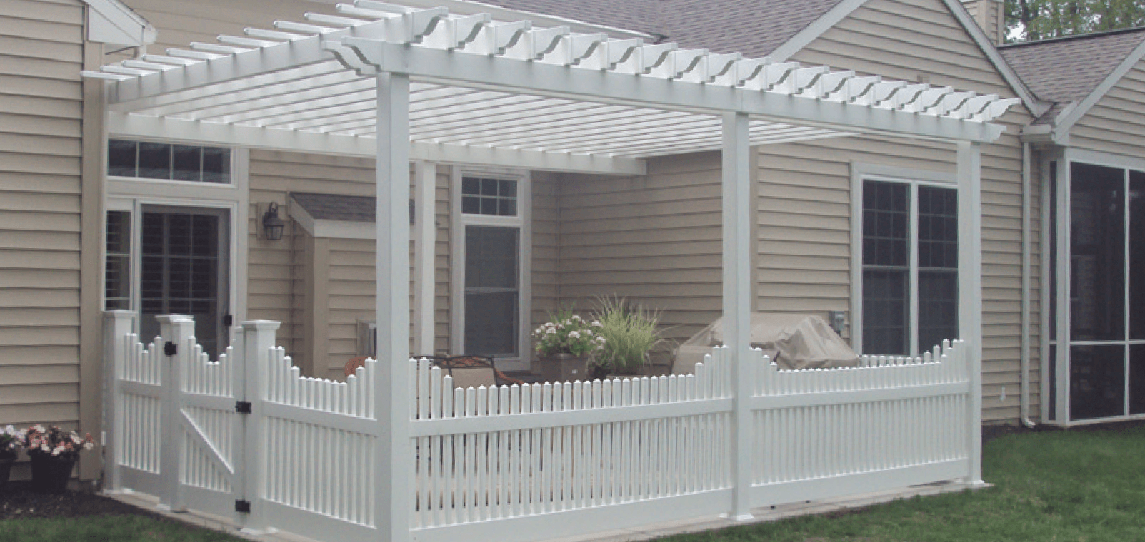 White pergola over a patio enclosed by a white picket fence, next to a house with tan siding.