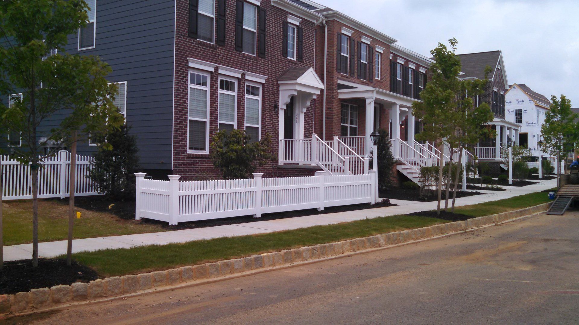 Row of townhouses with white picket fences and landscaping along a sidewalk.