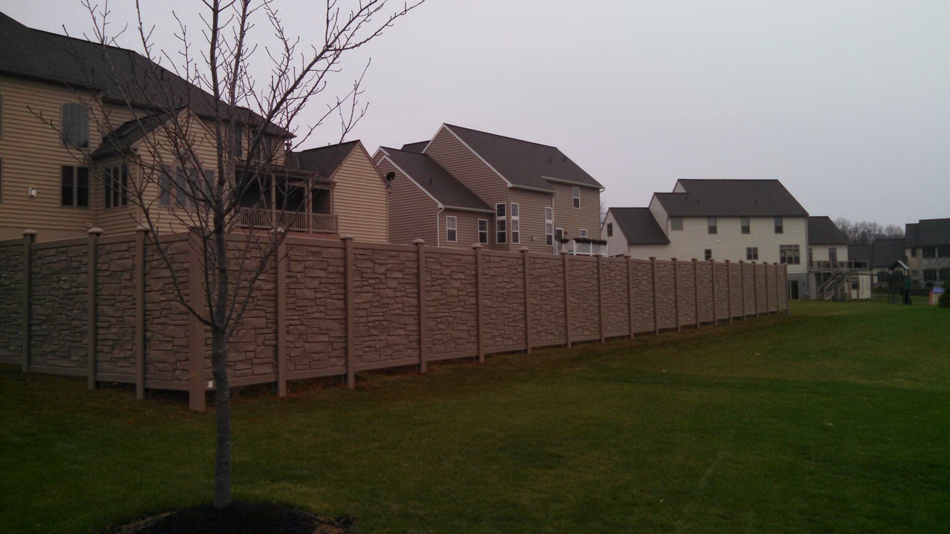 Long, textured brown fence bordering green backyard with houses in the background on a cloudy day.