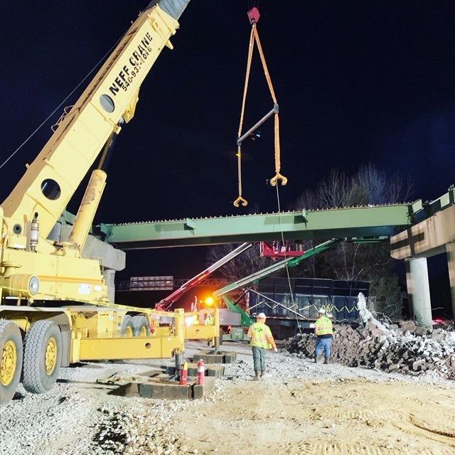 Crane lifting a damaged green bridge beam at night as workers inspect the repair site below