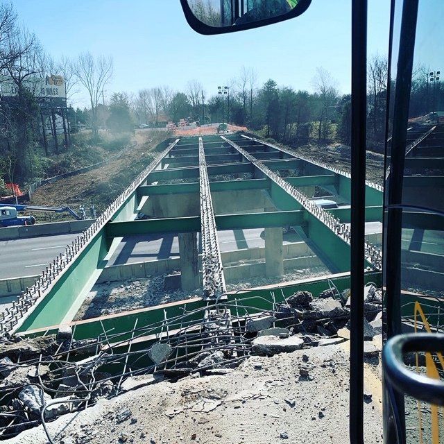 Collapsed bridge construction site viewed from a vehicle, with damaged green steel beams and debris below