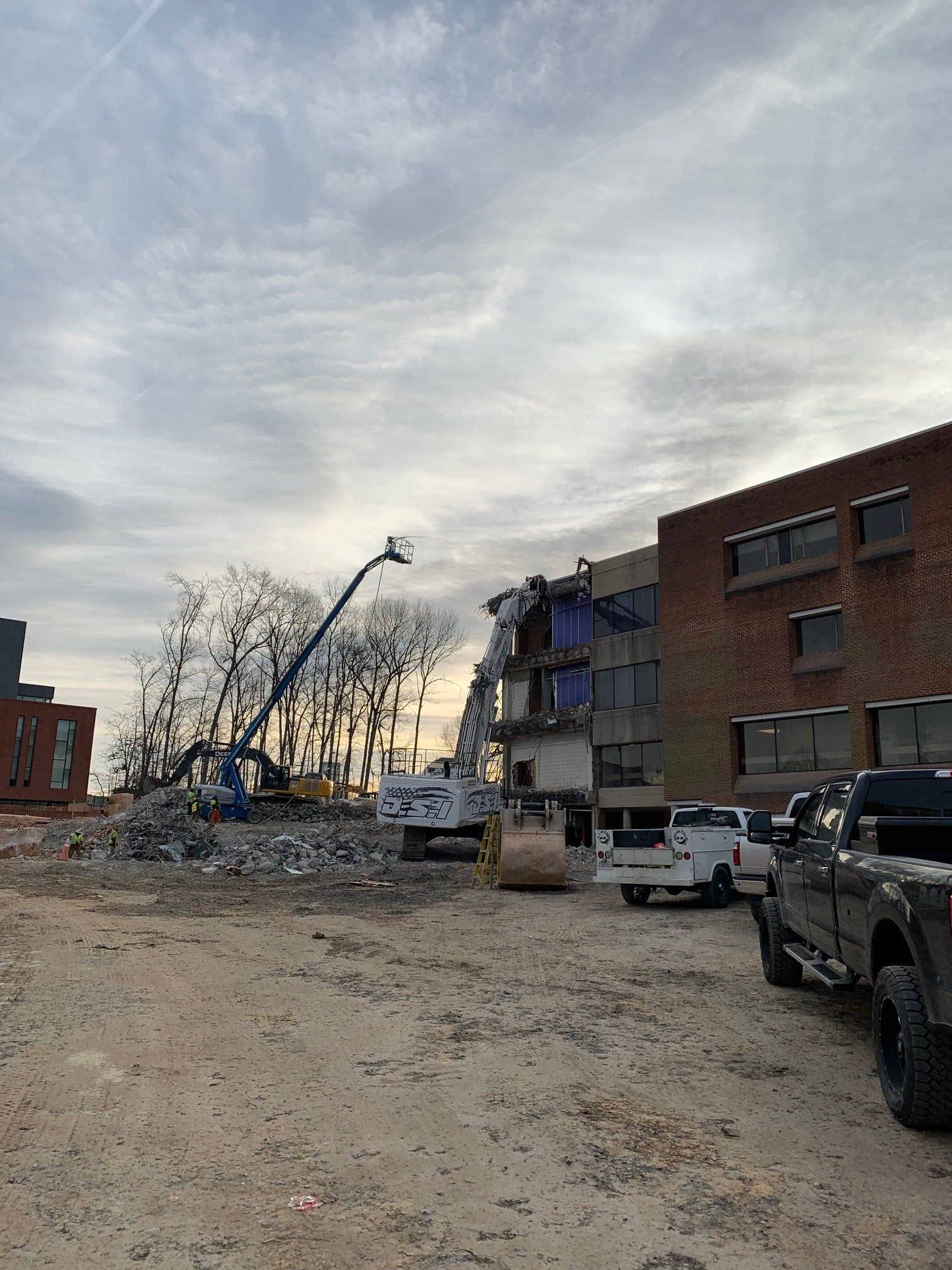 Damaged street beside a brick building, with rubble, a tilted utility pole, and parked trucks under a cloudy sky