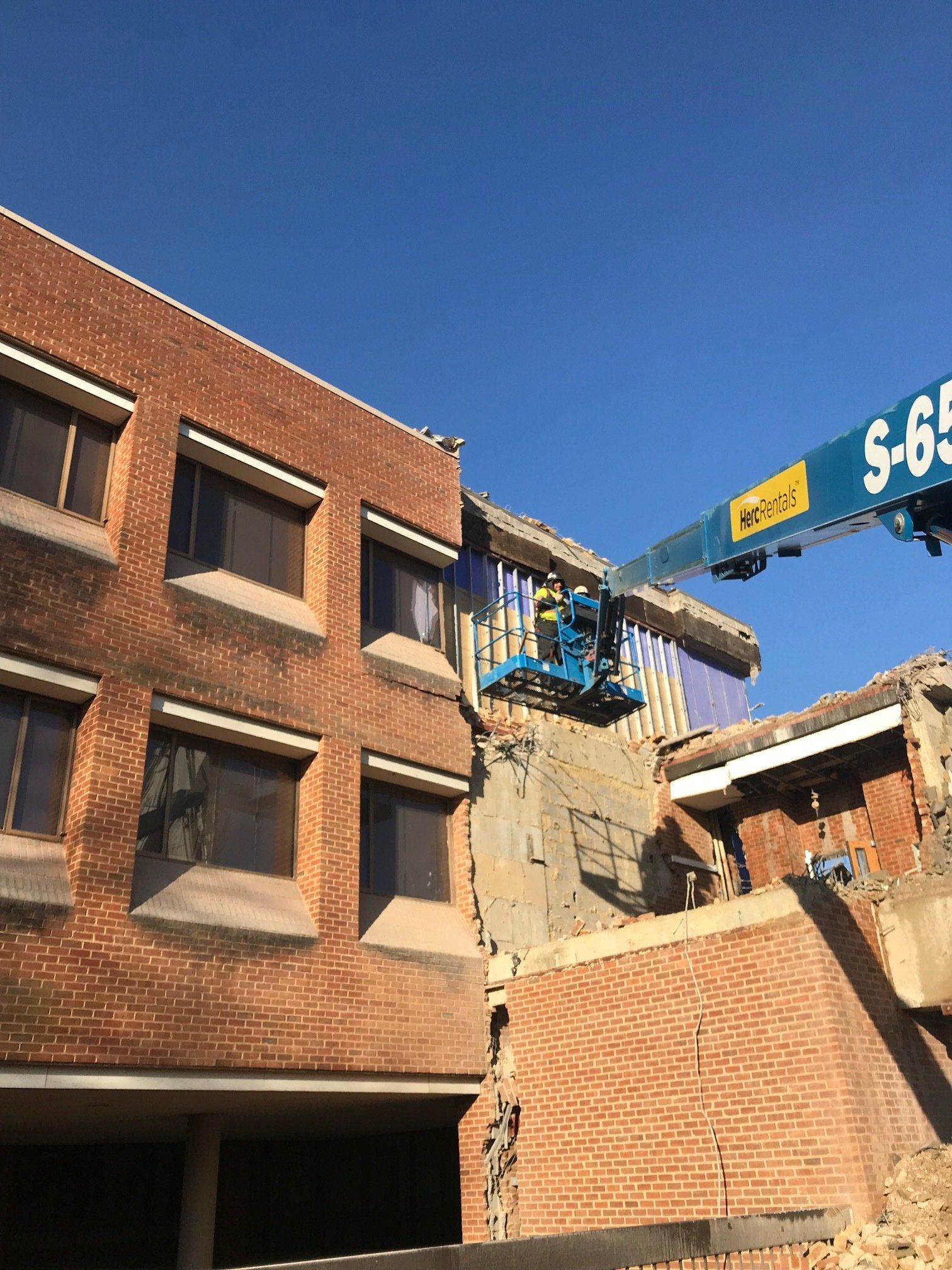 Brick building being demolished by a crane with debris falling under a clear blue sky