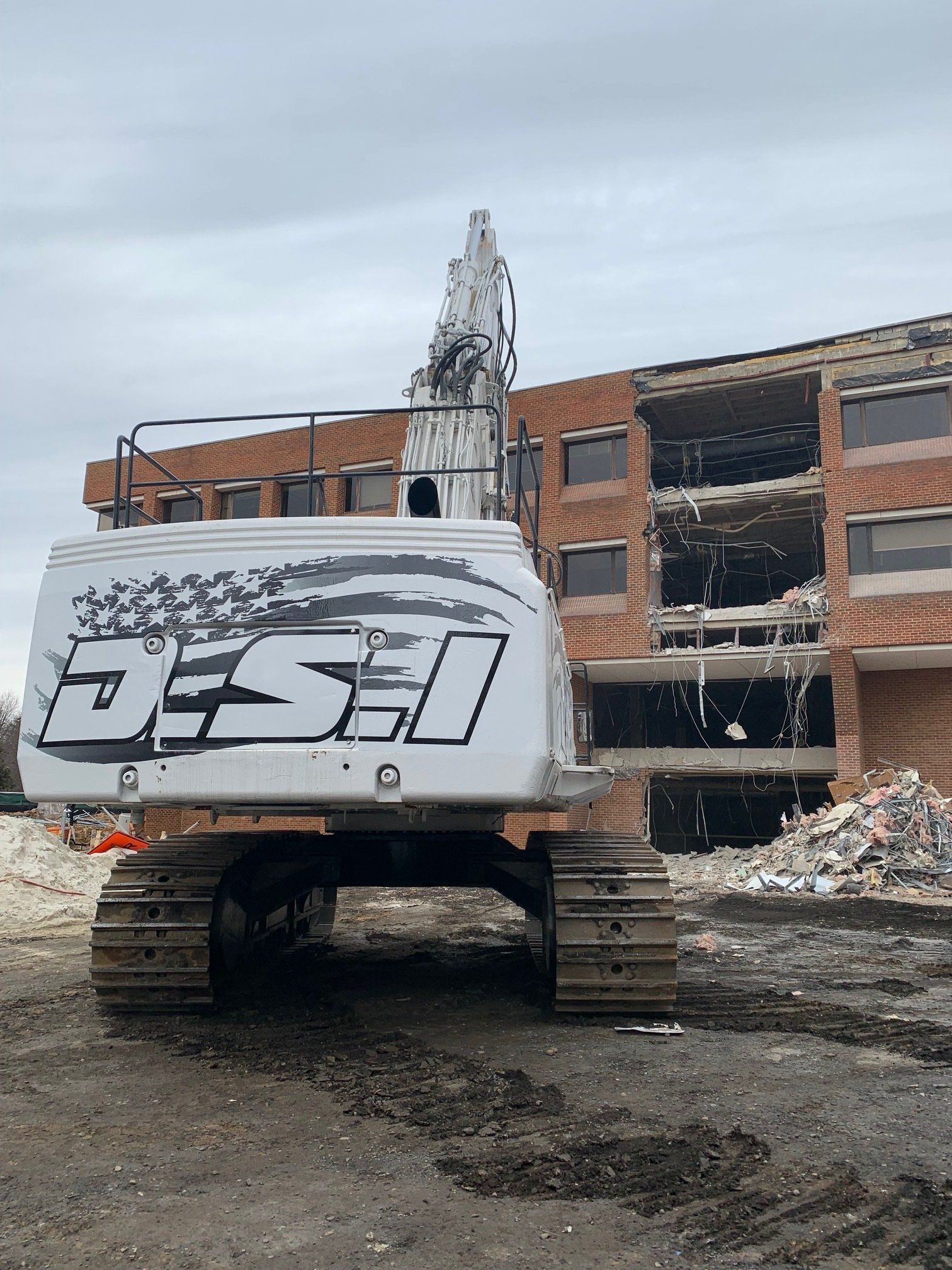 Tracked excavator in front of a partially demolished brick building under an overcast sky