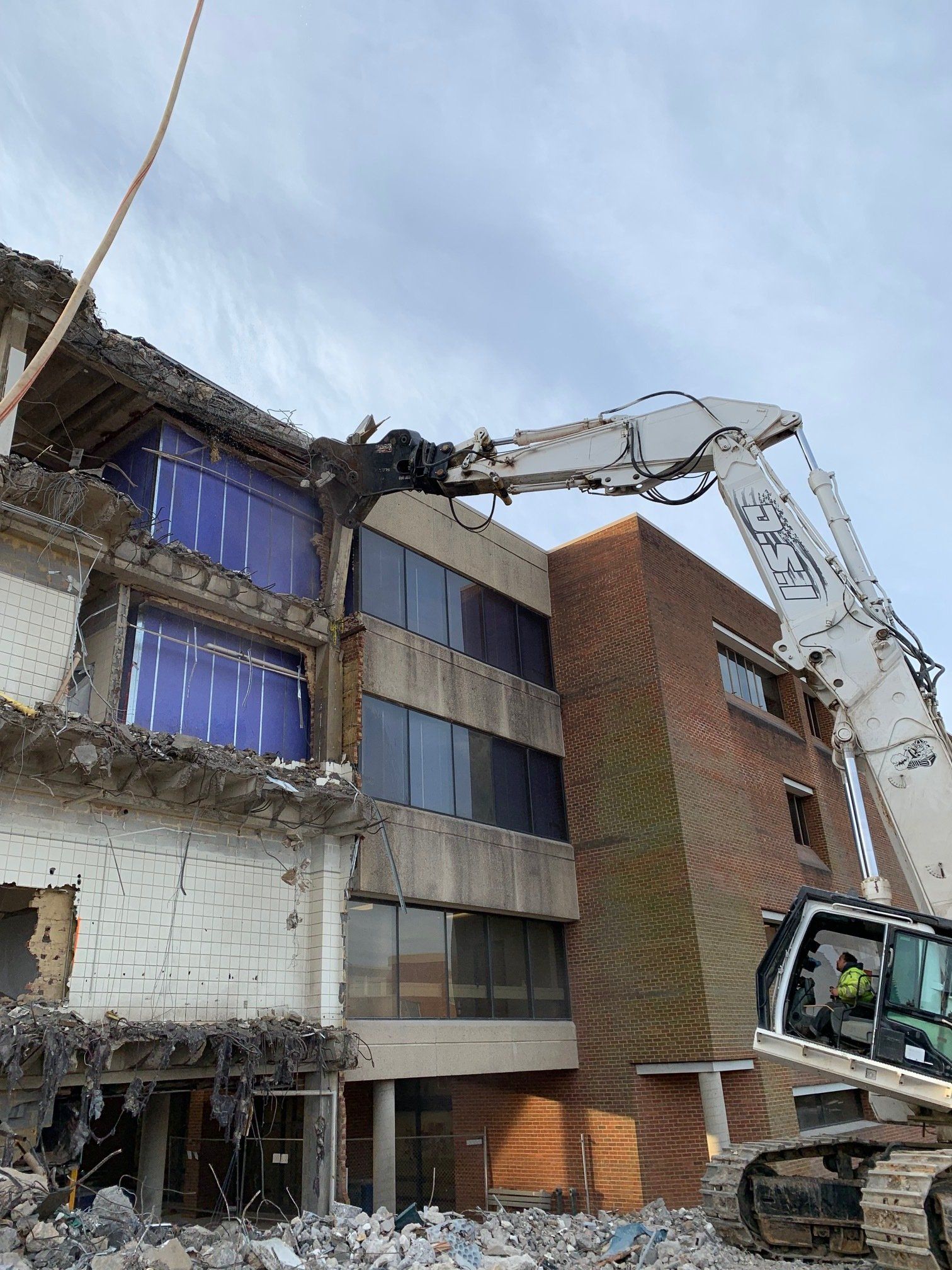 Excavator demolishing a damaged multi-story building under a cloudy sky