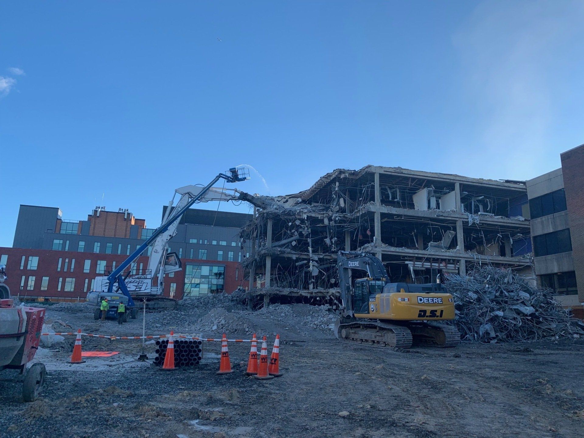 Demolition site with a partially collapsed building, excavator, debris, and orange cones under a blue sky