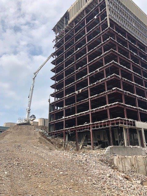Partially constructed steel-frame high-rise with a concrete pump and rubble foreground under a cloudy sky
