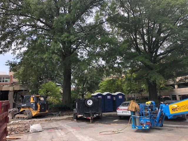 Construction equipment and dumpsters under large trees at a work site, with a white car nearby.