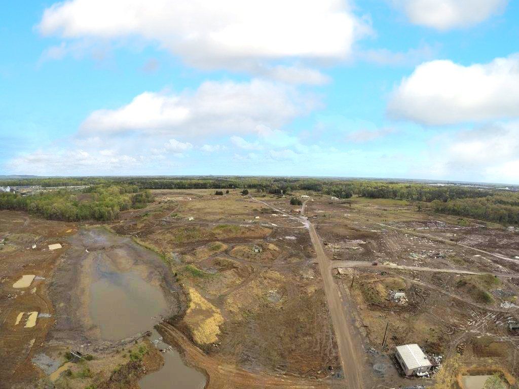 Aerial view of a barren landscape with muddy water pits, dirt roads, and a small white vehicle under a cloudy sky