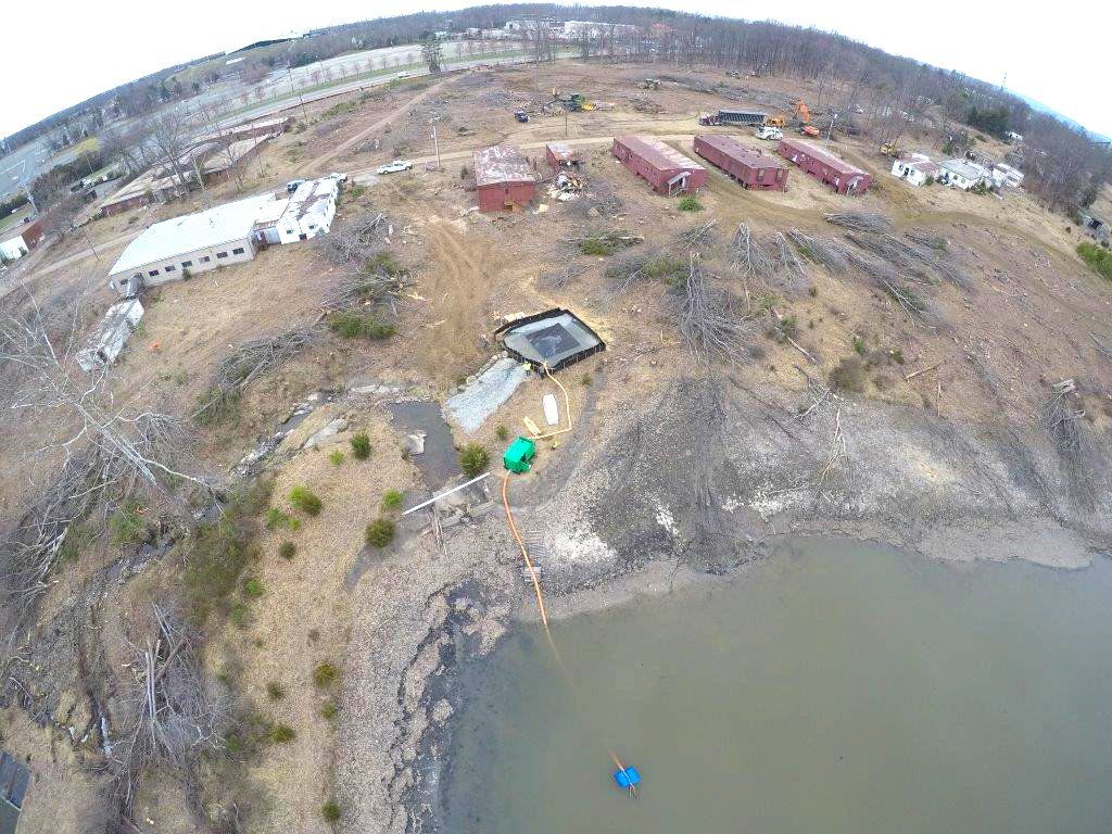 Aerial view of a muddy lakeshore with a dock, small structures, and buildings in the background.