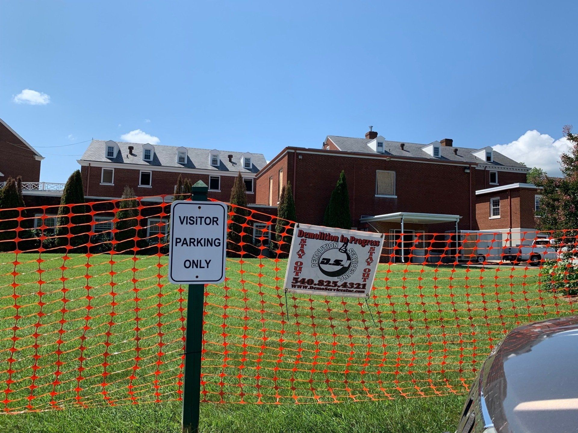 Orange construction fencing in front of brick buildings and signs on a grassy lot under a blue sky.