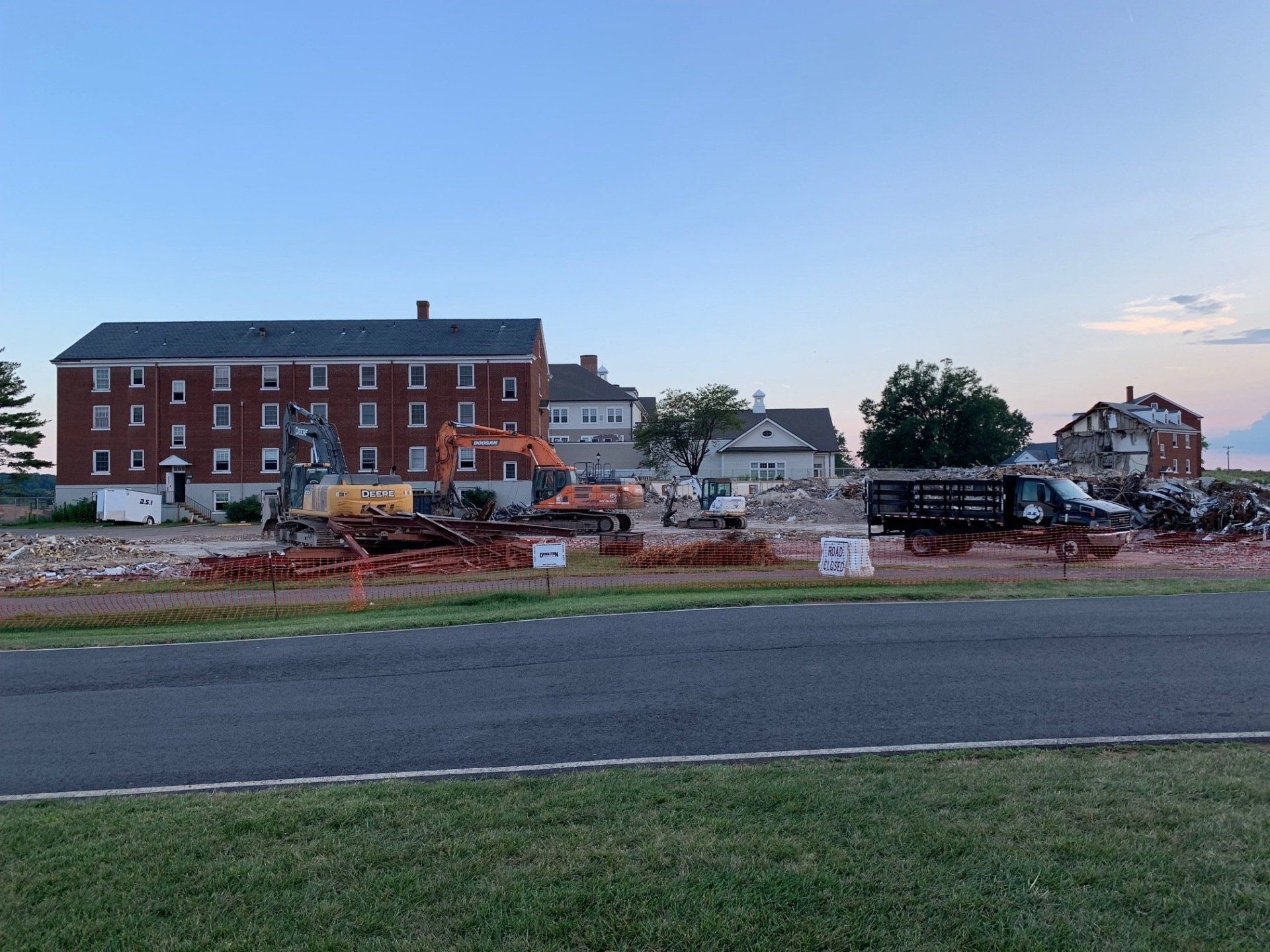 Roadside construction site with excavators, rubble, and a red brick building at dusk